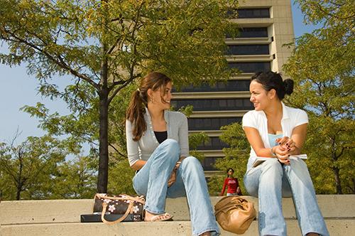 Two students sitting on steps talking and laughing with campous buildings in background.